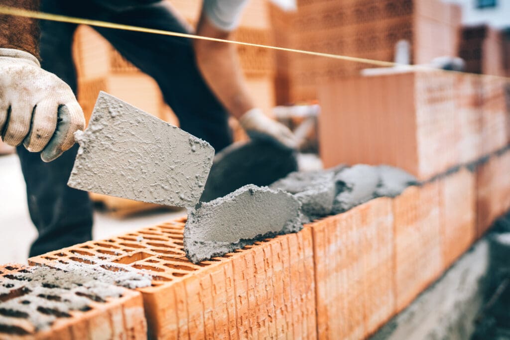 A construction worker spreads mortar on bricks with a trowel while building a brick wall.