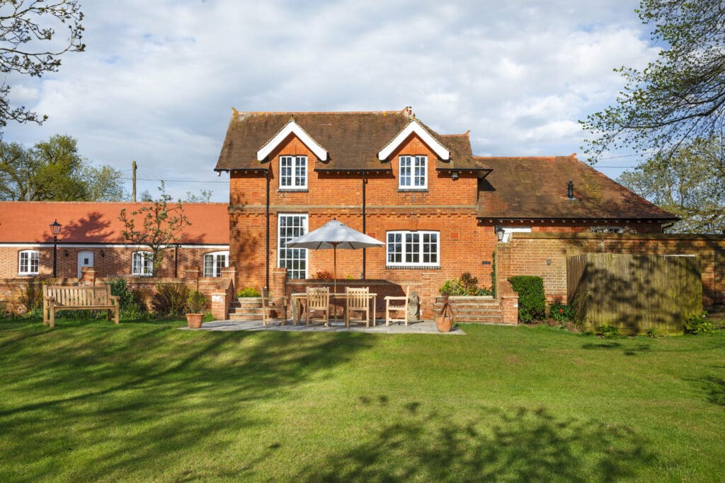 A red brick house with white trim, a patio with wooden furniture and an umbrella, and a large green lawn under a partly cloudy sky.