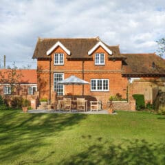 A red brick house with white trim, a patio with wooden furniture and an umbrella, and a large green lawn under a partly cloudy sky.