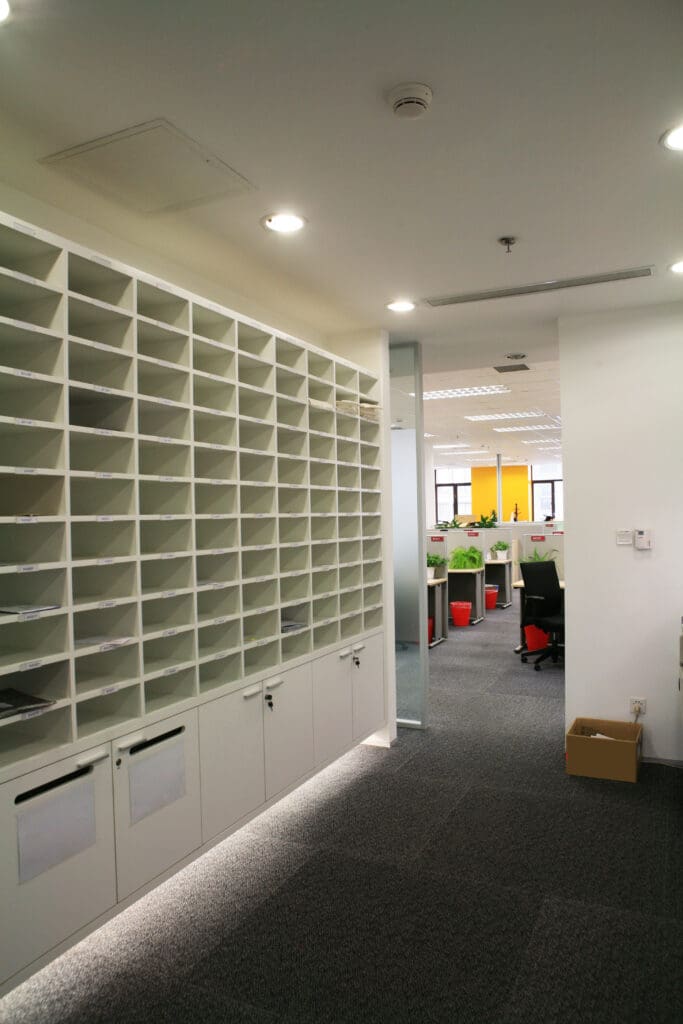 A modern office mailroom with empty cubby shelves, a few storage cabinets below, and an open doorway leading to a workspace with desks and chairs.
