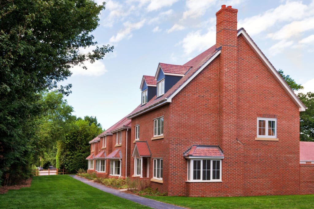 Large red-brick house with multiple chimneys and dormer windows, set on a green lawn with trees and a pathway leading to the entrance.