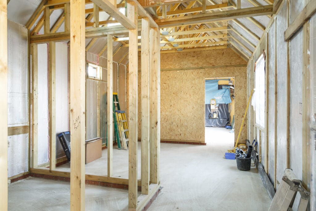 Interior of a house under construction with exposed wooden framing, insulation, and building materials scattered on the floor.