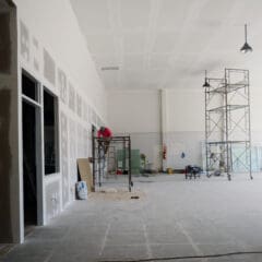 Large indoor construction site with scaffolding, unfinished walls, exposed drywall, and workers performing installation or repairs.