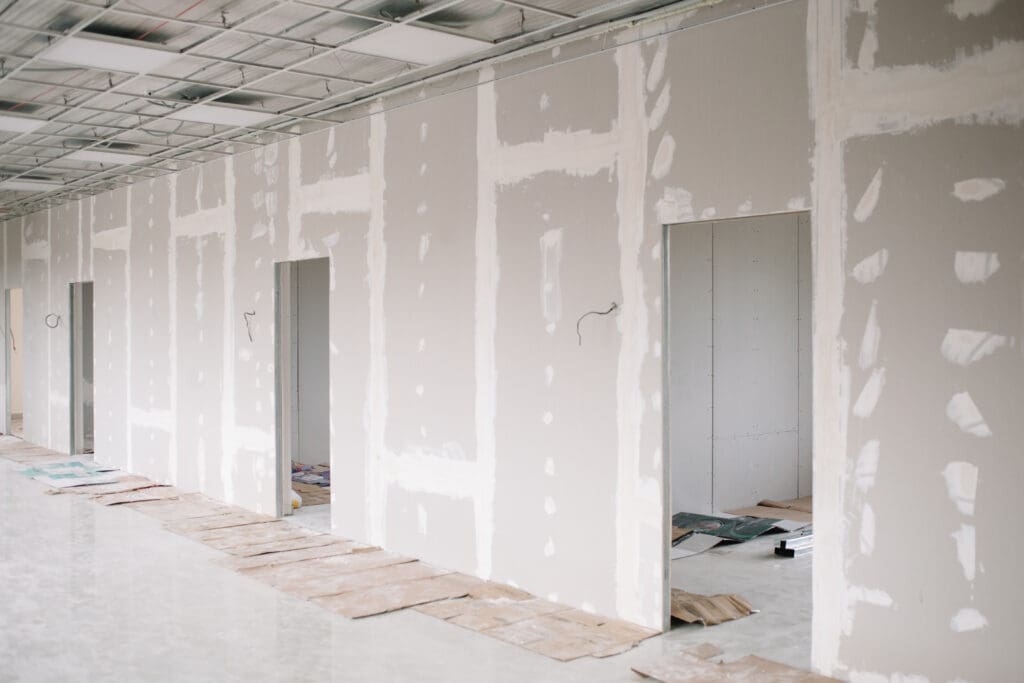 Unfinished interior wall with drywall panels, exposed seams, and multiple door openings under construction in a building.