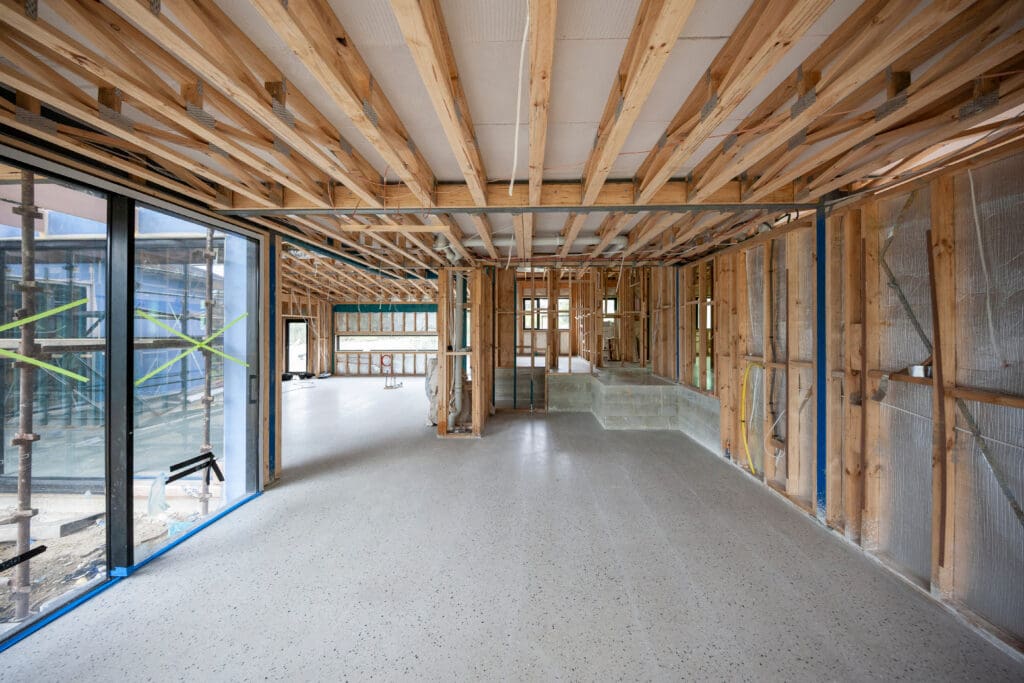 Large room under construction with exposed wooden framing, unfinished walls, and glass windows. Concrete floor is complete; visible scaffolding and construction materials outside.