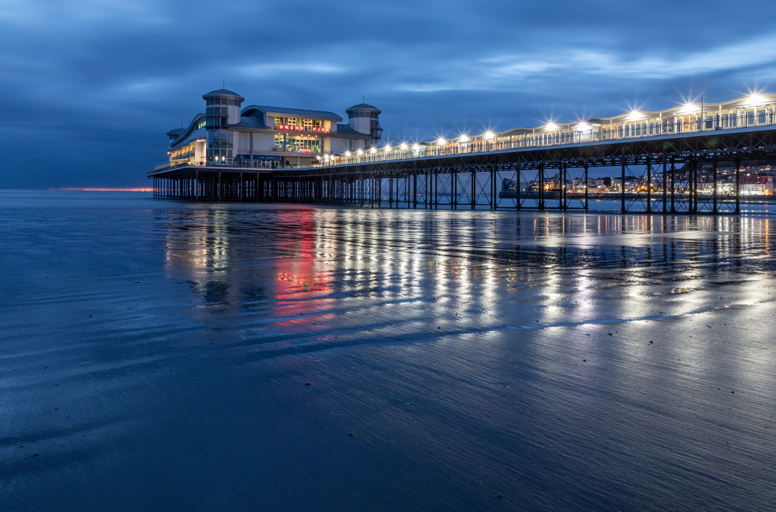 A lit pier extends over the sea at dusk, with reflections of lights shimmering on the wet sand and calm water beneath a cloudy blue sky.