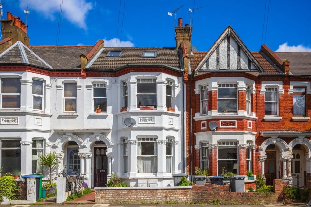 Row of Victorian-style terraced houses with bay windows, brick and stucco facades, and small front gardens under a blue sky.