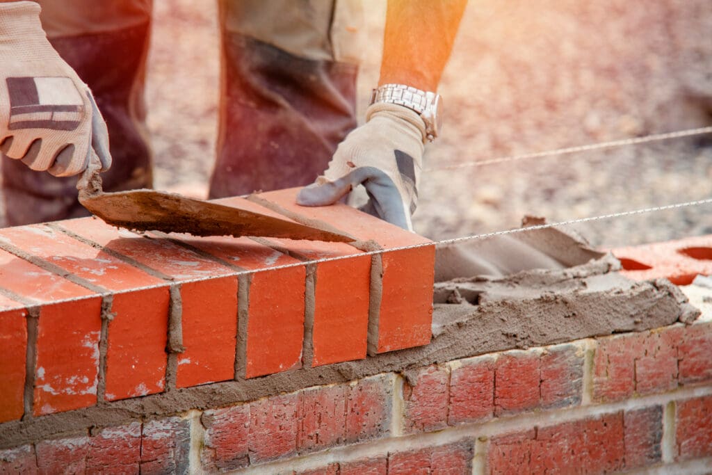 A person wearing gloves uses a trowel to lay red bricks and mortar while constructing a brick wall.