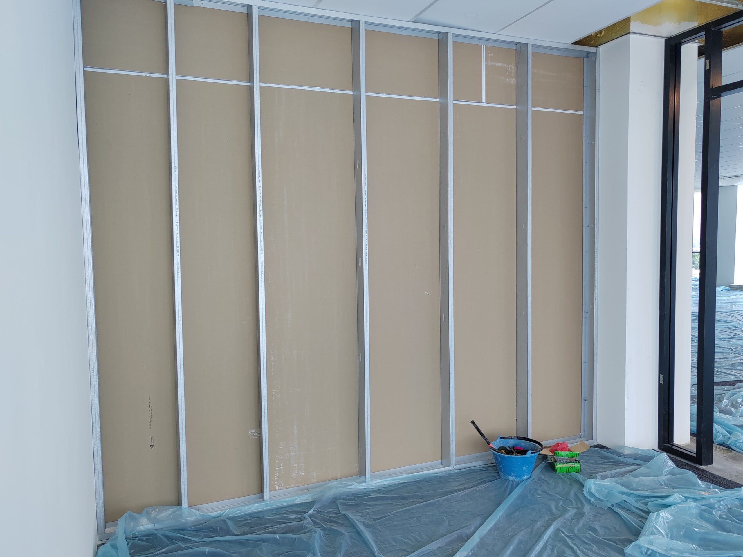 A partially finished drywall partition with exposed metal studs, plastic sheeting on the floor, and a blue bucket with tools and paint supplies nearby.