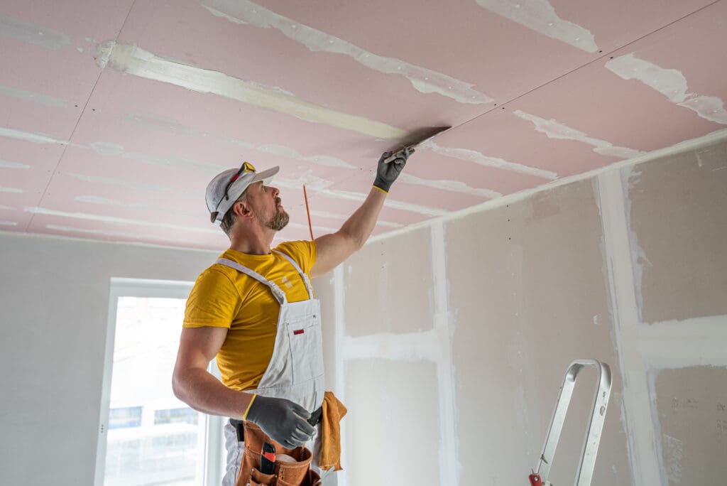 A worker in overalls applies joint compound to seams on a drywall ceiling in a room under construction.