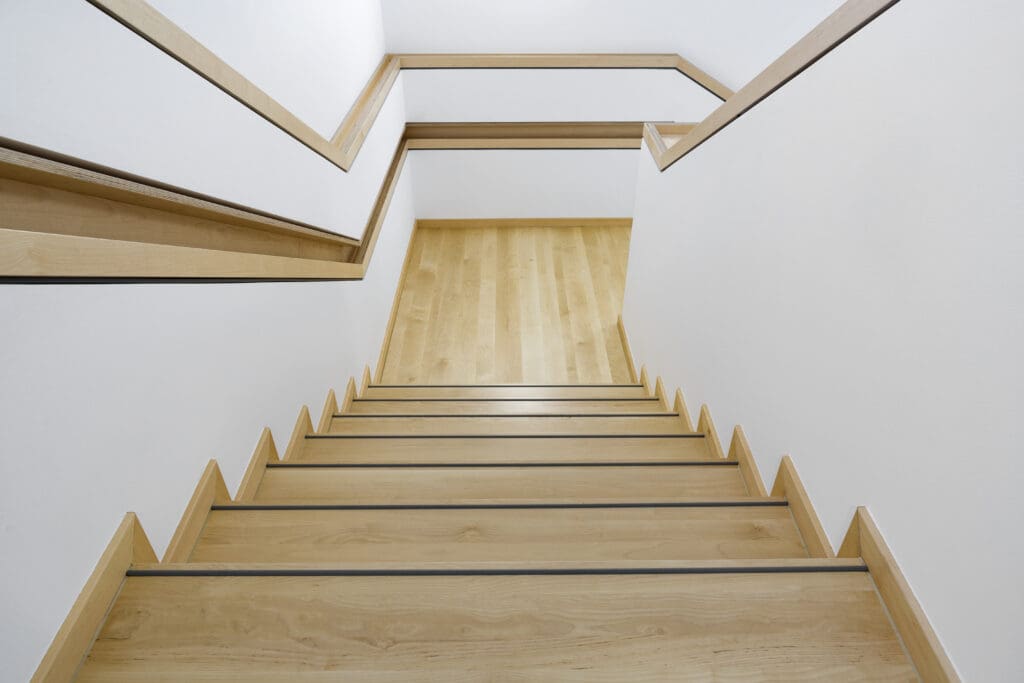 View looking down a modern wooden staircase with metal edging and white walls, featuring simple, minimalist design and clean lines.