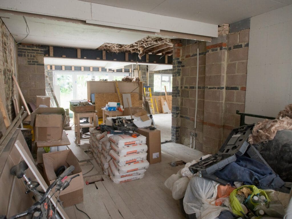 Interior of a house under renovation with exposed brick walls, construction materials, tools, and stacks of plaster bags scattered throughout the unfinished space.