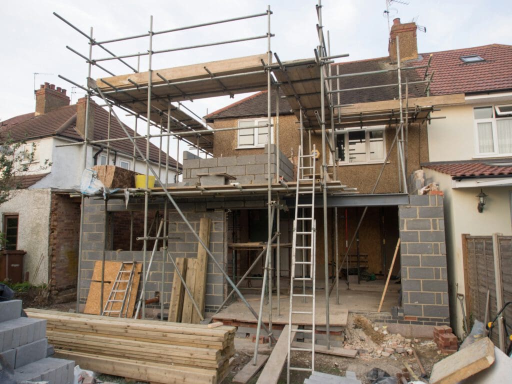 A two-story house under construction with scaffolding, building materials, and ladders in the front yard. The extension is partially built with exposed brickwork and wood panels.