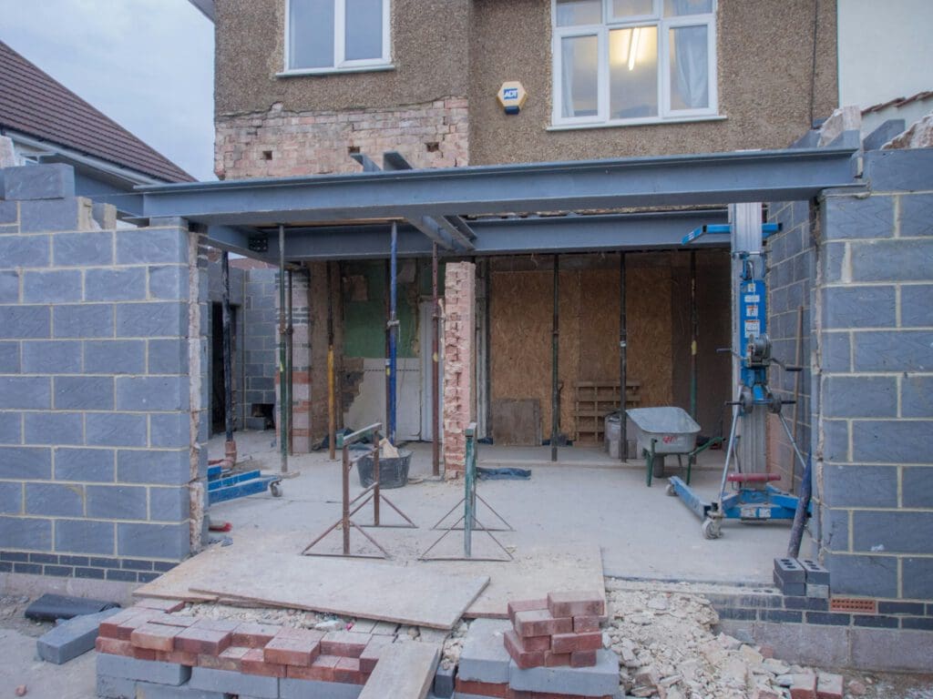 Partially constructed house extension with exposed brickwork, steel beams, construction equipment, and stacks of bricks in the foreground.