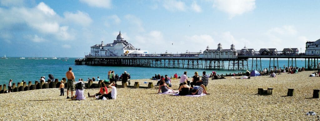 People sit and relax on a pebble beach near a pier extending into the sea under a partly cloudy sky.