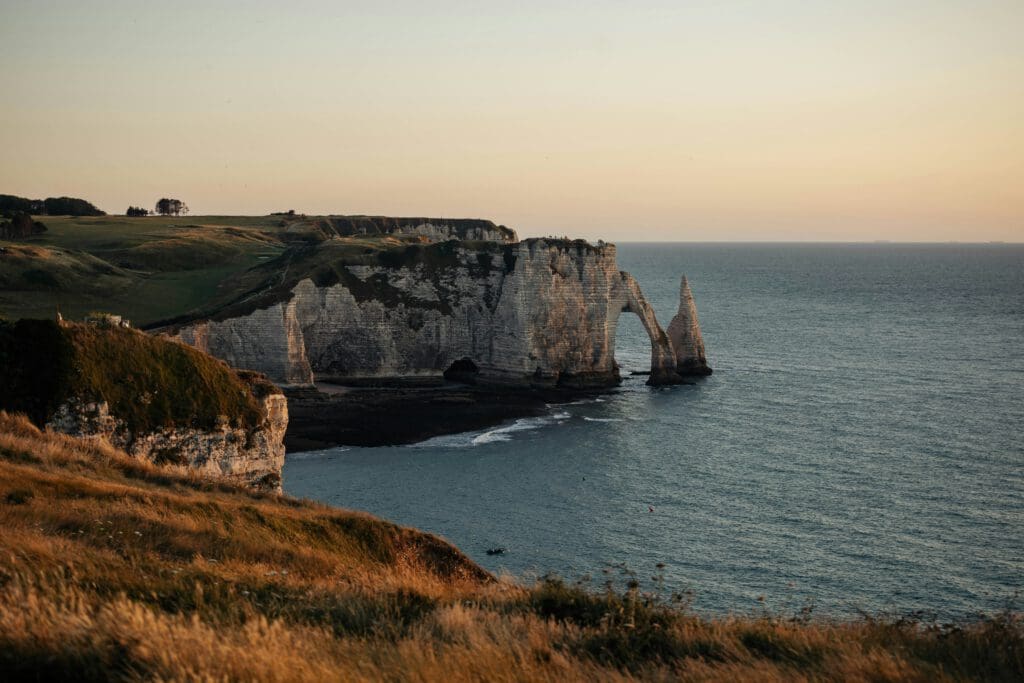 Cliffs with natural rock arches overlook the sea at sunset, with grassy fields in the foreground and calm water below.